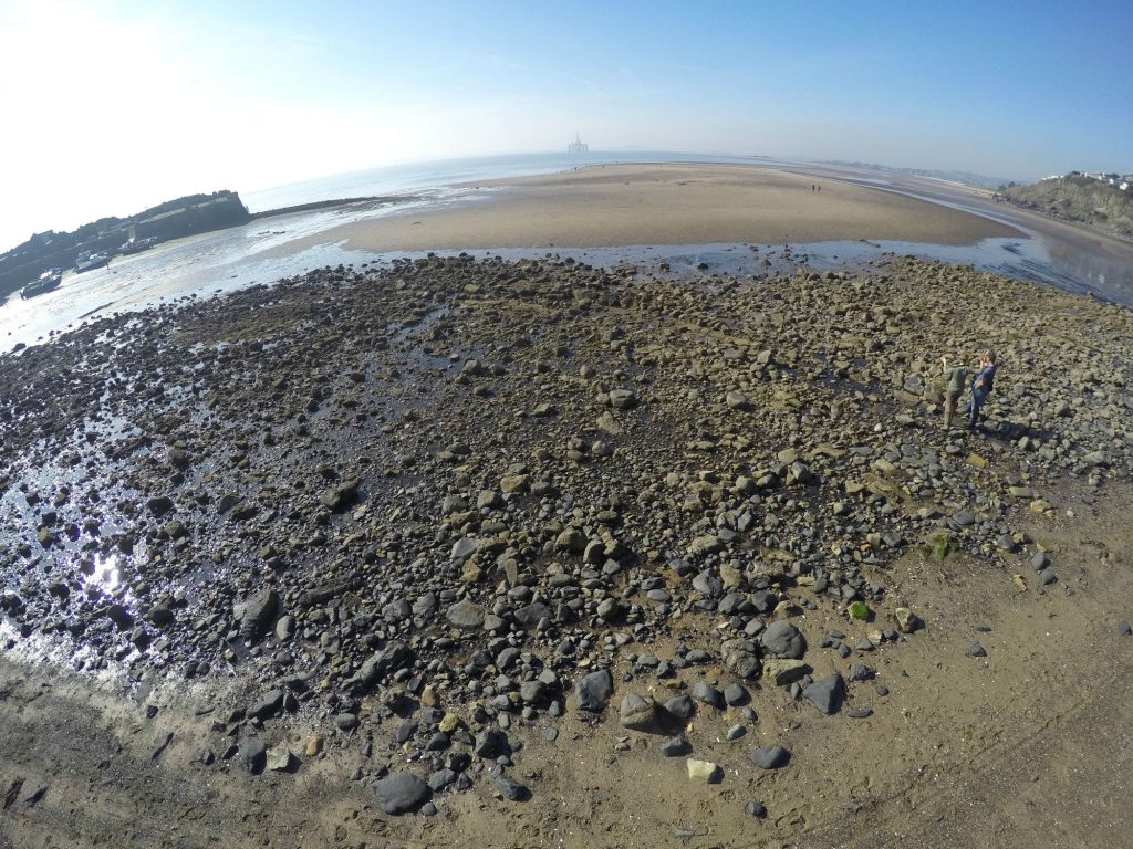 An aerial fisheye view of a sandy beach with the tide out and a large spread of stone in the foreground. A straight line of flat stones runs diagonally across the spread