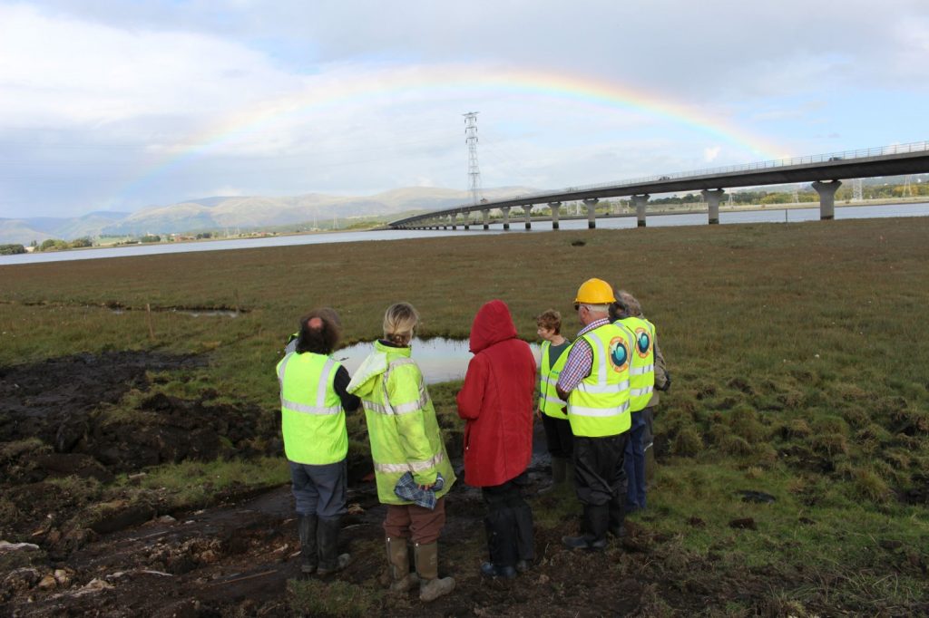 A group of people in hi-vis clothing standing on grass looking across an expanse of intertidal saltmarsh towards a distant river channel, with a rainbow on the opposite bank
