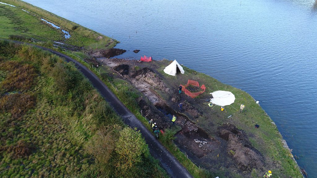 An aerial view of an archaeological trench on a grassy riverbank at high tide with water up to a stone sea wall
