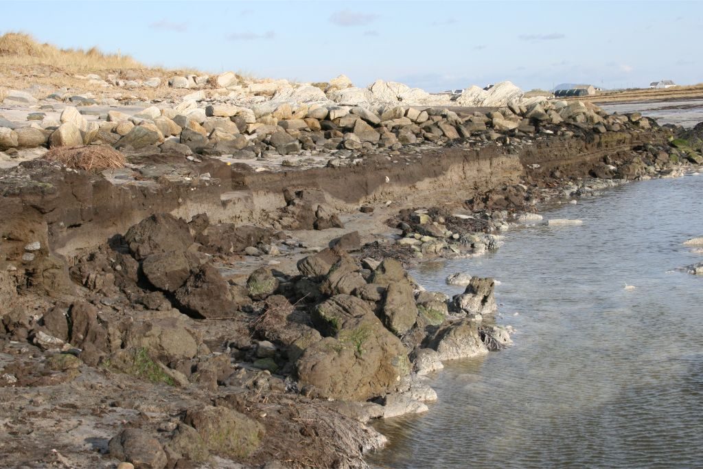 A beach with a vertical eroding face at the waters edge, and a low spread stone wall on the shore behind
