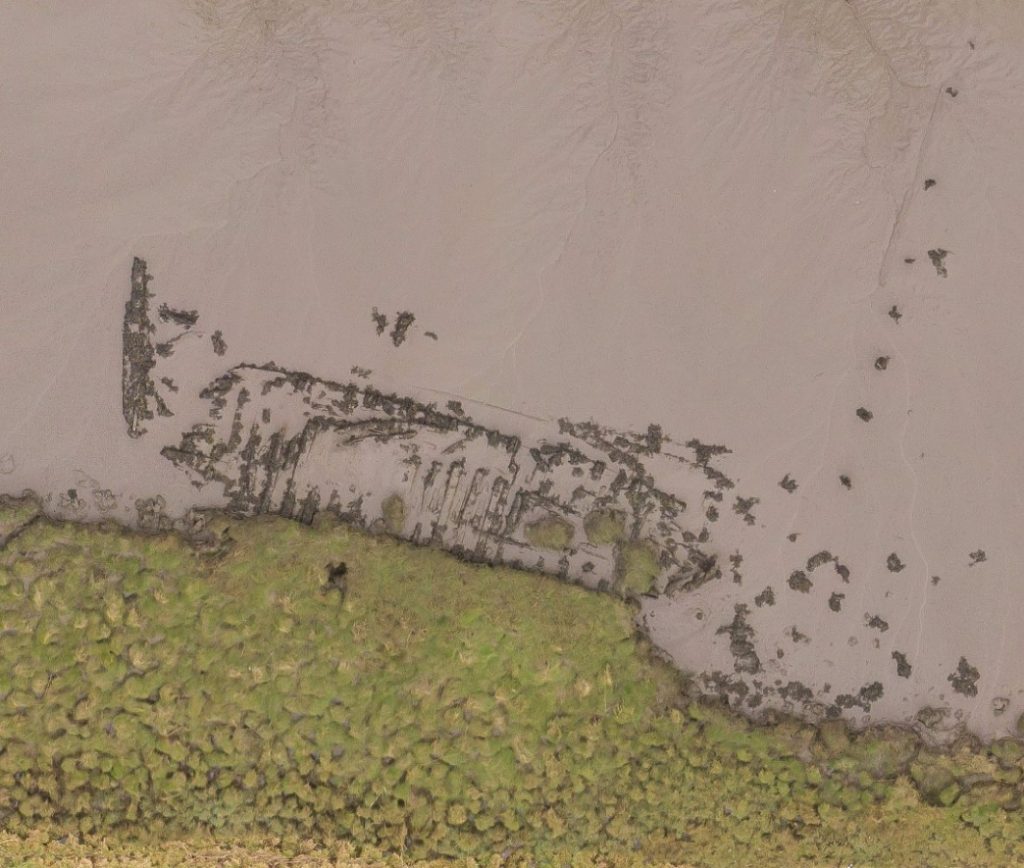 An aerial photograph of the outline of a wrecked wooden boat mostly buried in mud on a riverbank