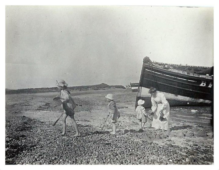 A black and white photo of three small children in summer clothes playing on a beach with large wooden boats visible on the shore behind them