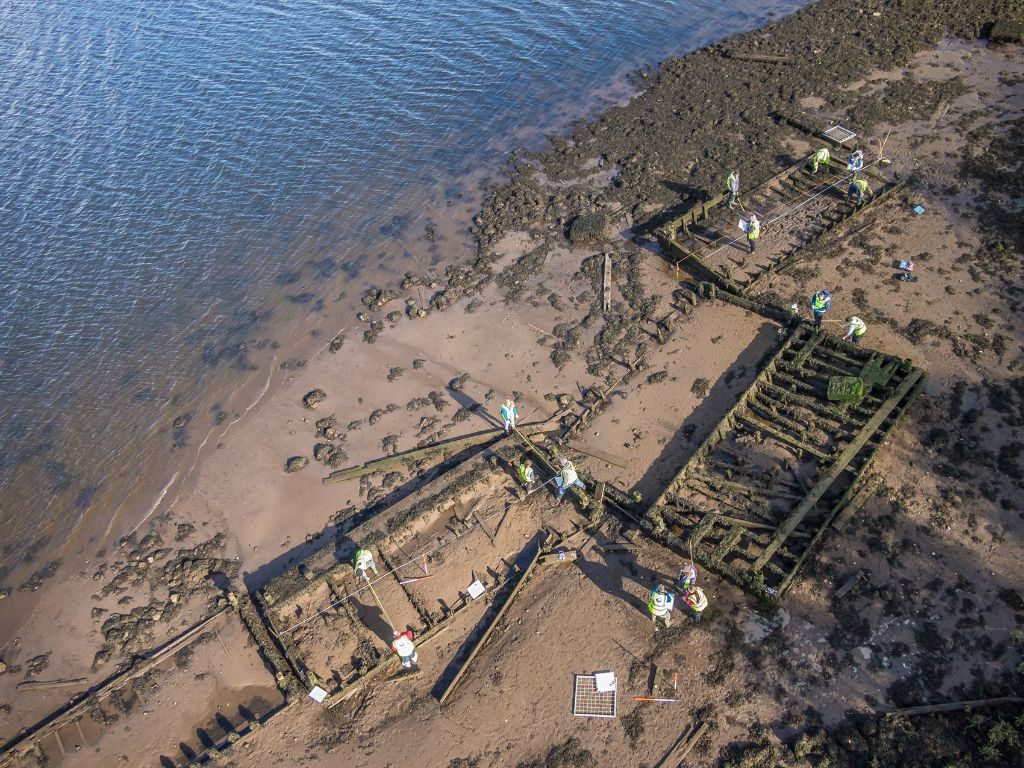 An aerial view of a group of dilapidated rectangular wooden structures half-buried in mud on the waterfront with a group of people wearing hi-vis vests dispersed around them, taking measurements and writing on clipboards