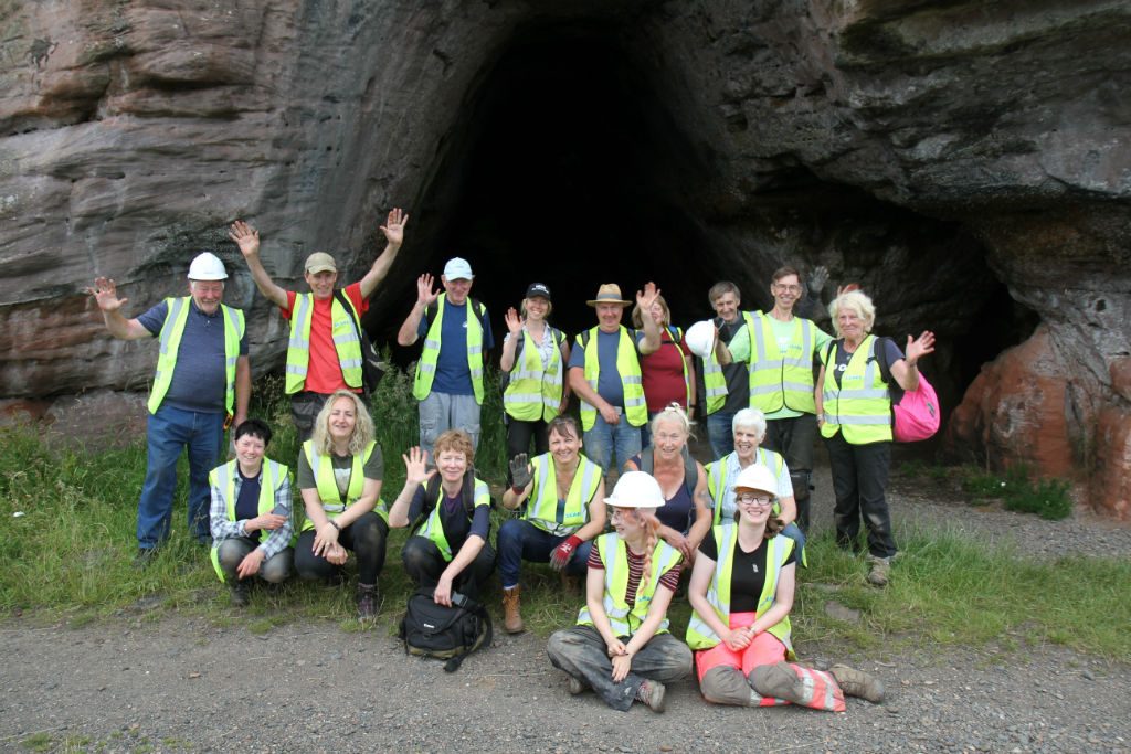 Group of people wearing high vis vests, standing and kneeling in front of a cave, smiling and waving at the camera