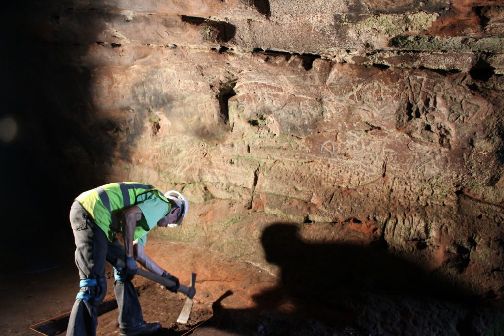 Man wearing hard hat and high vis vest mattocking in front of a cave wall bearing ancient carvings