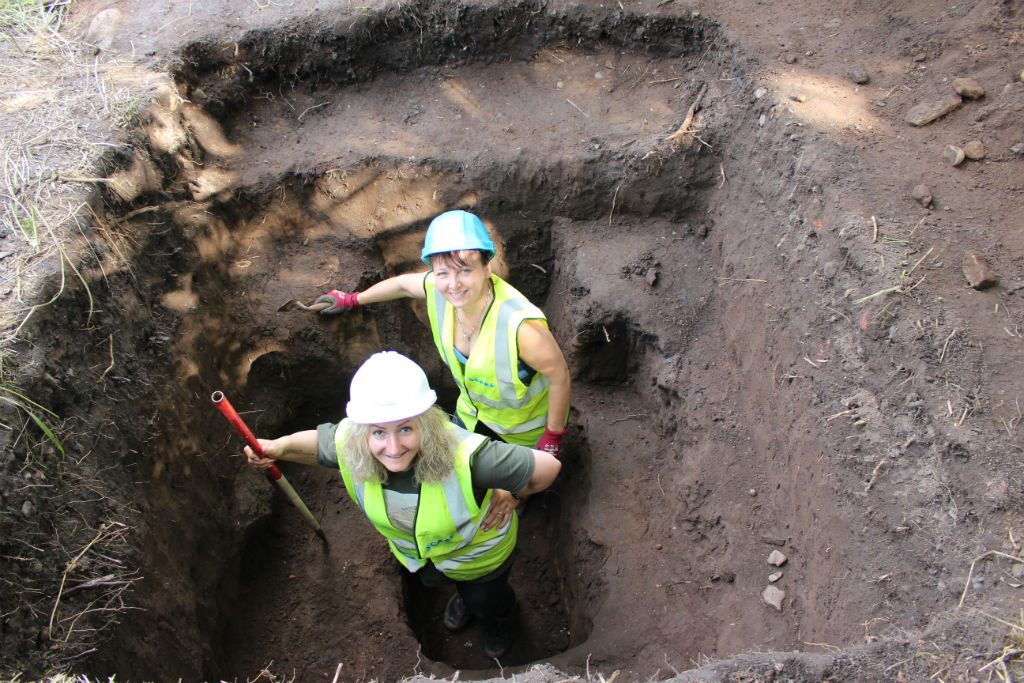 Two women wearing hard hats and high vis vests standing in an archaeological test pit smiling up at the camera