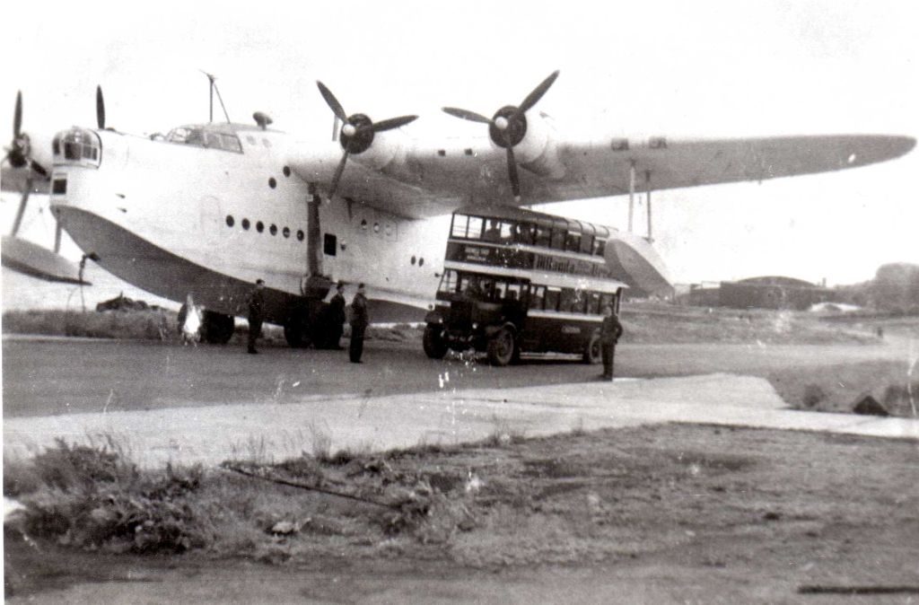 An old black and white photo of a huge flying boat being towed up a road, with an old-fashioned double-decker bus passing underneath one of its wings