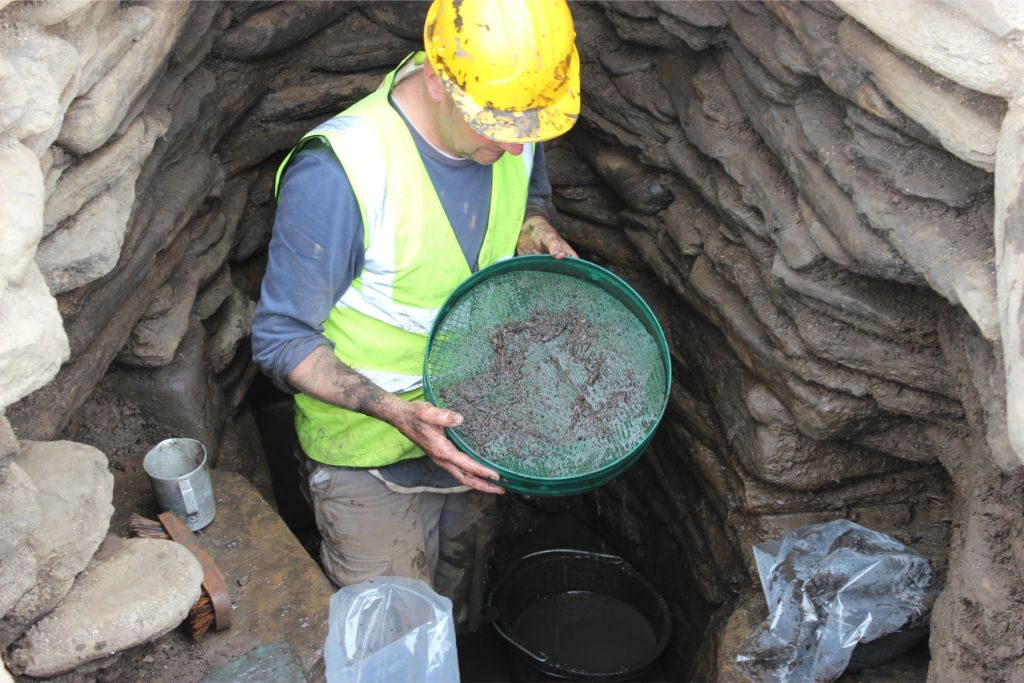 A man wearing a hard hat and hi-vis standing in an underground drystone structure holding a large seive which contains fragments of plants and other organic material 