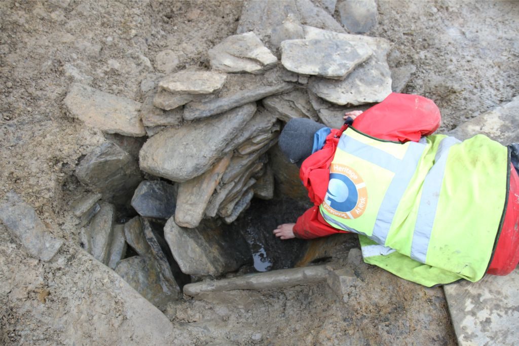 A person wearing a hi-vis vest bending down to reach into a small, stone-lined hole in the ground