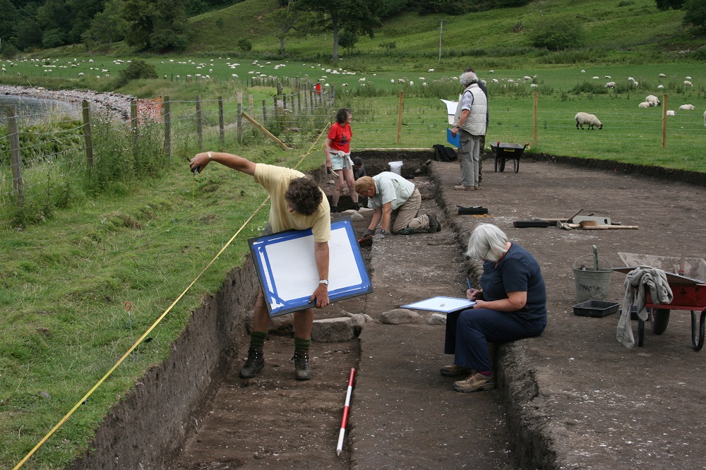 An archaeological trench, with people measuring and drawing in the foreground, and people with folders and paperwork in the background
