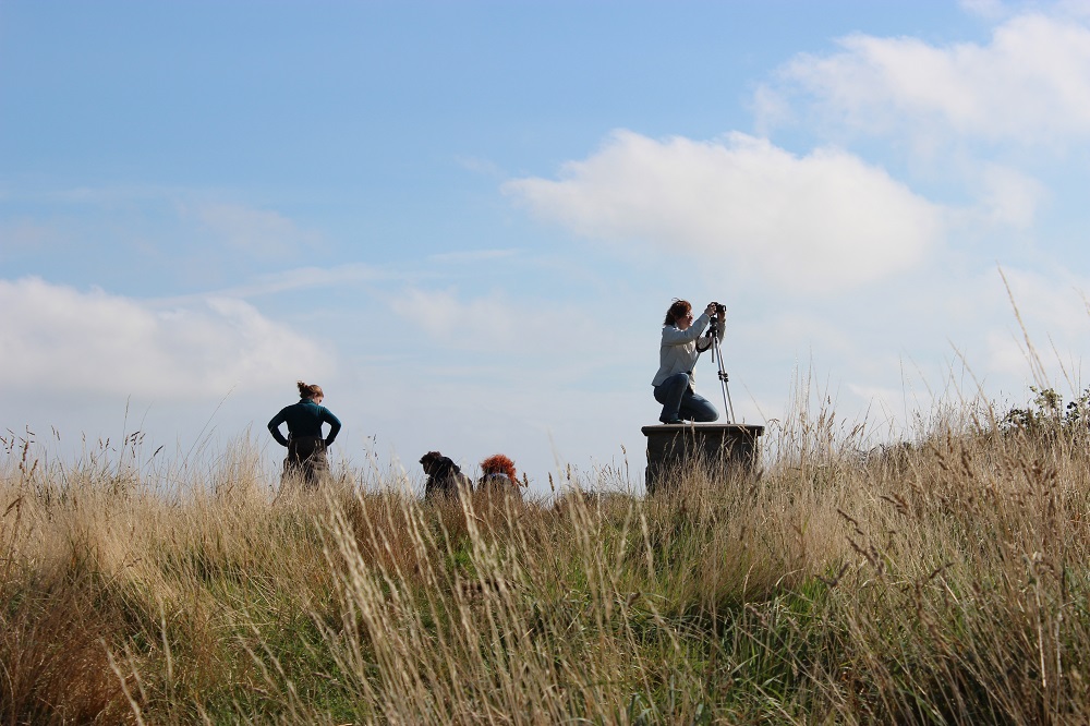 A grassy hill with a group of people on the horizon, one filming with a camera on a tripod