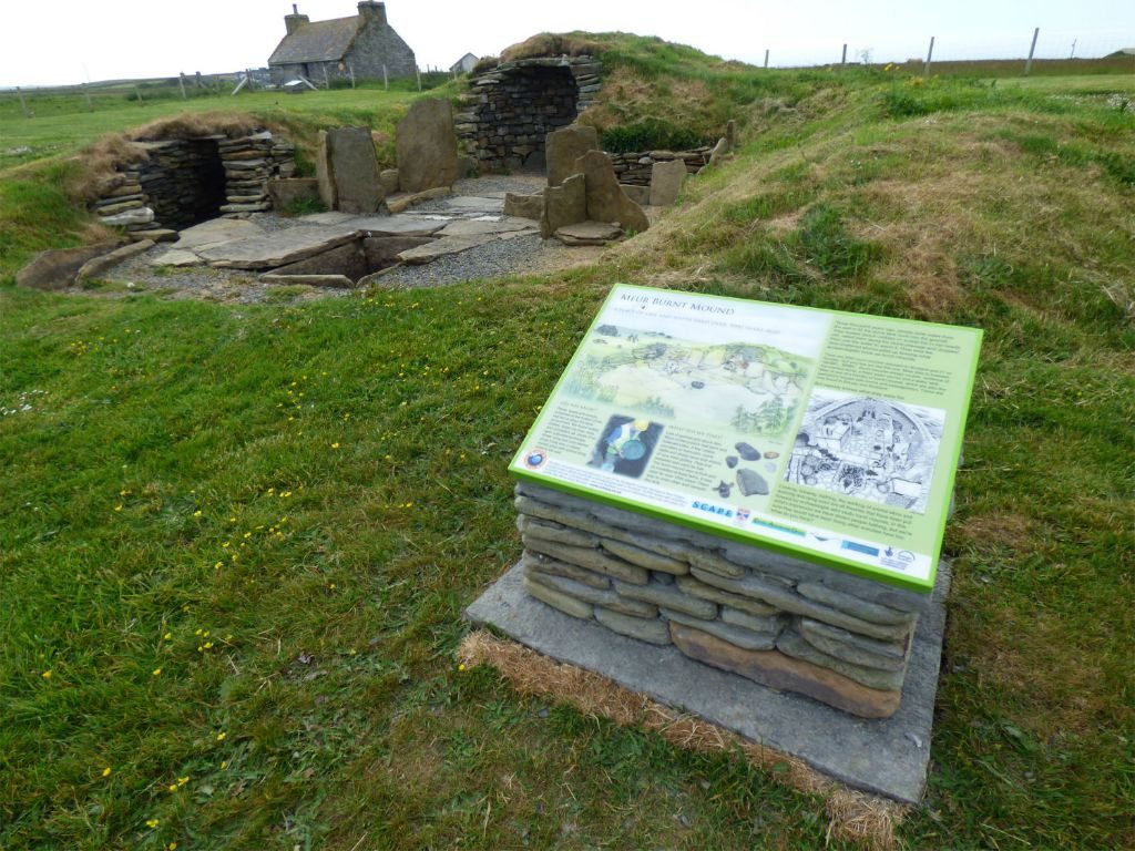 Stone structures built of large upright flagstones forming small compartments with corbelled drystone structures, capped with turf, in a grassy area with a large interpretation board in the forground