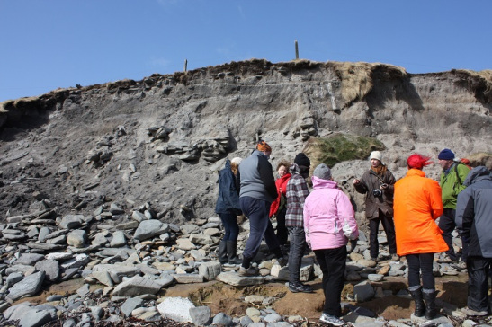 A group of people standing on a pebble beach below an eroded edge containing stonework and different soil deposits