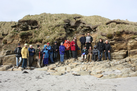 A group of people standing on a beach in front a cliff with the remains of a broch on top