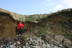 A girl standing next to an eroding coast edge, with a pit cut into the surface and flat stones visible lining the pit.