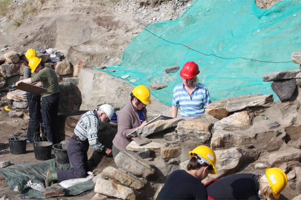 A group of people on an archaeoloical site, drawing and trowelling, surrounded by stone walls and sand