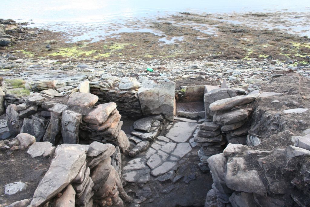 An archaeological site of drystone structures, paved stone surfaces and upright slabs, on a pebble beach with the sea behind