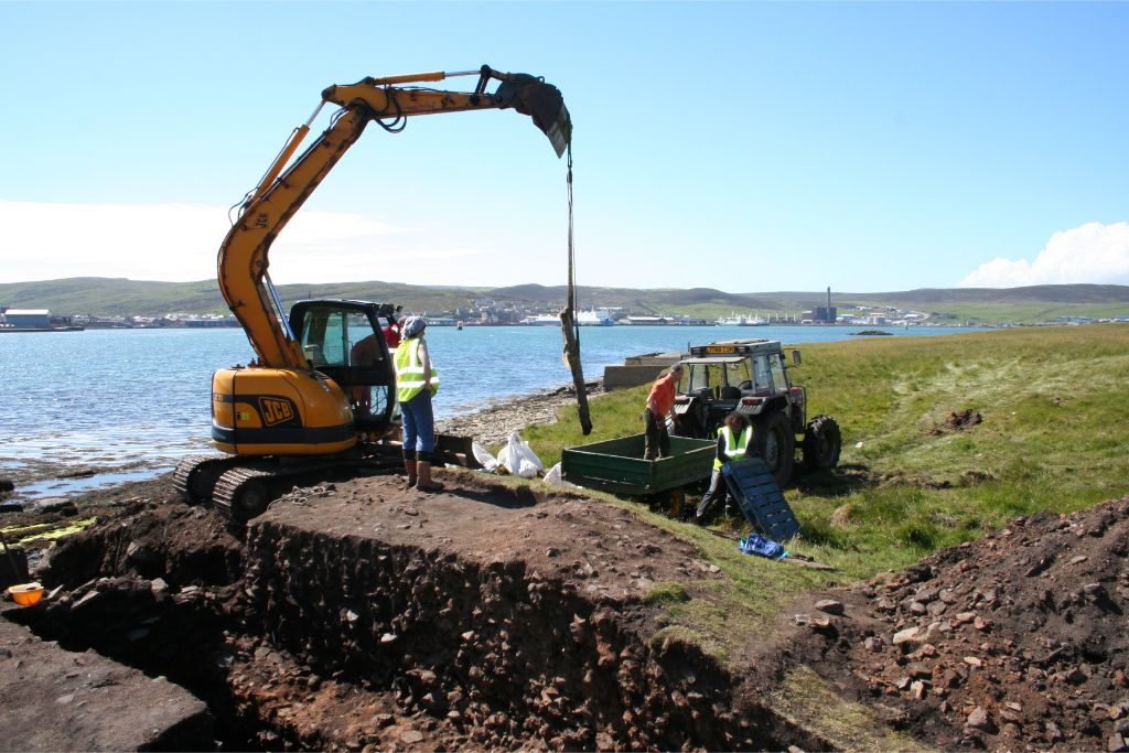 An archaeological trench next to the sea, with a digger lifting a large stone slab onto a trailer and trailer
