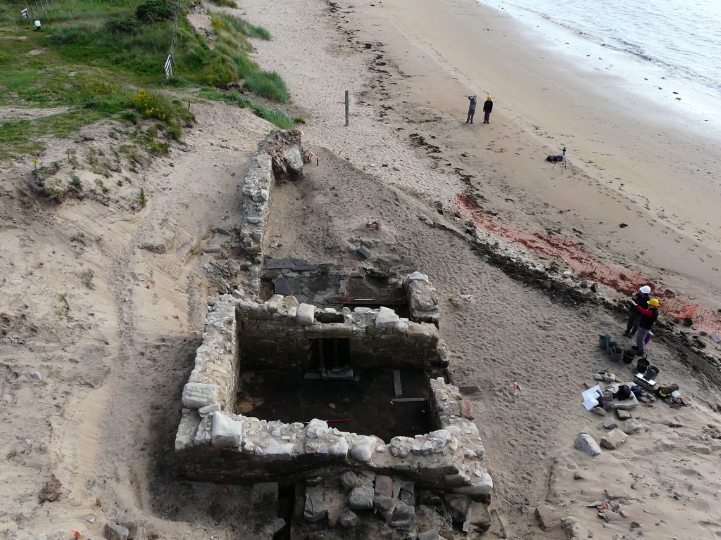 Overhead view of a sandy beach with a ruined rectangular stone building exposed by the excavation of a sand dune, with one corner of the building destroyed