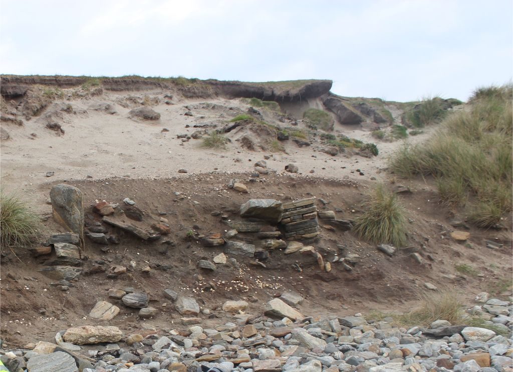 An eroding sandy cliff face on a beach with stone walls and dark layers of soil visible in the section