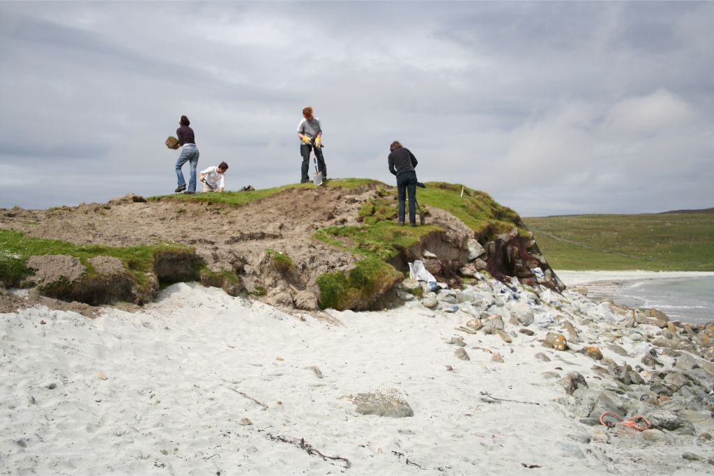 A grass-covered mound on a sandy beach, with stones protruding from the face, and people standing on the top, removing squares of turf