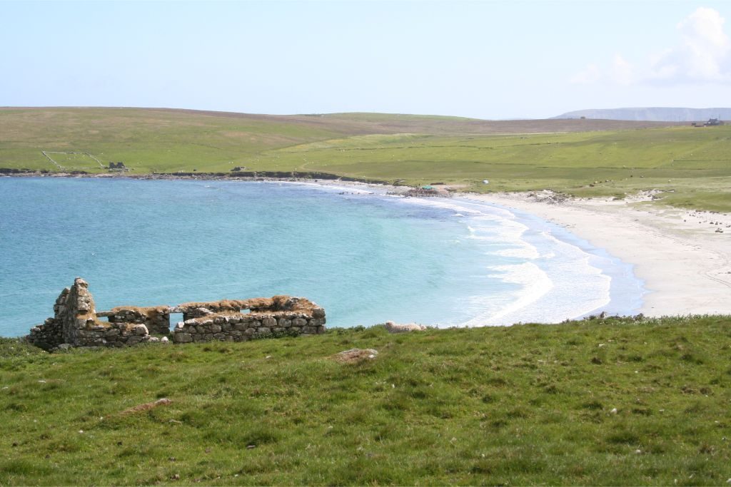 A sweeping sandy beach with grassy slope behind and a ruined stone building overlooking the bay