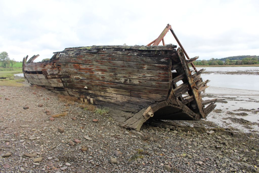 A wooden boat sitting on a mud and pebble riverbank, resting on its starboard side. The planking around the transom and part of the planking of the port side of the hull is gone