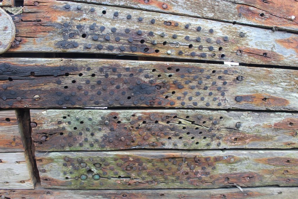 A close up shot of wooden planking of the hull with a grid of small round holes, some filled with wooden pegs