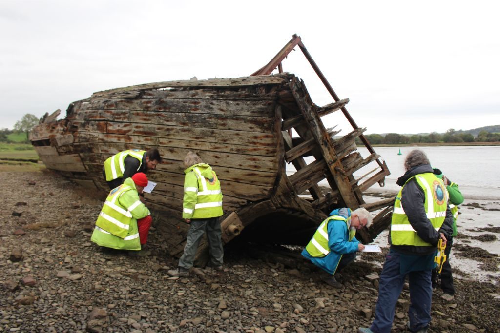 The same wooden boat on the riverbank, with a group of people in hi-vis vests clustered around the stern recording the vessel writing notes and holding tape measures