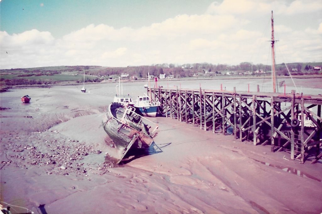 A faded, old photograph of a riverbank with a wooden boat hauled up on the muddy bank. The boat is the same as the other photos, but looks intact,The name FAUNA is visible on the name plate on the transom