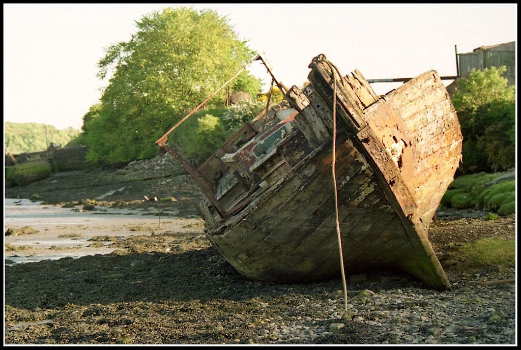 A wooden boat sitting high up on a beach, the view shows the bow and front end, with metal plating around the stem at the front. The iron plating has partly fallen off, the paintwork is peeling and the planking looks slightly rooten. The boat has clearly not been used for some time, but is intact