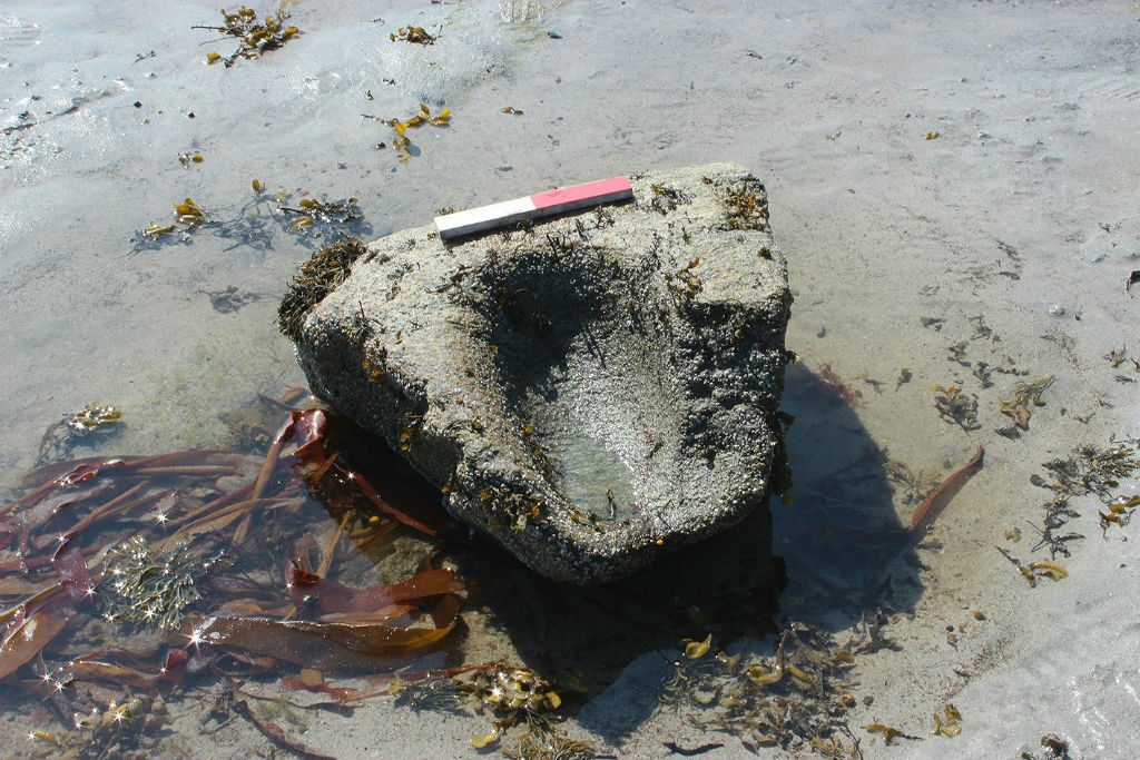 Large stone with smooth hollow carved into its surface lying upon sand wiht shallow water and seaweed around it