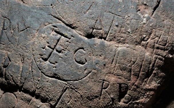 Close up of cross with embellished arms inside a circle surrounded by initials - all carved into a sandstone cave wall